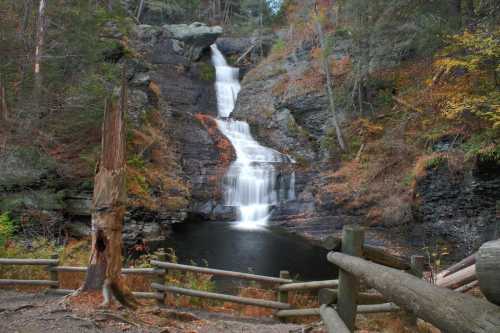 A serene waterfall cascades down rocky cliffs into a calm pool, surrounded by autumn foliage and wooden fencing.