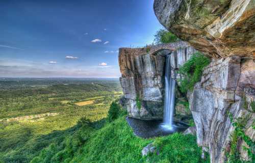 A stunning waterfall cascades from a rocky cliff, surrounded by lush greenery and a vast landscape under a clear blue sky.