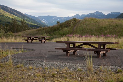 Two wooden picnic tables on a paved area surrounded by grass and mountains in the background under a cloudy sky.