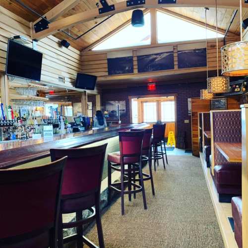 Interior of a rustic bar with wooden walls, high ceilings, and a long counter with stools. Bright natural light from windows.