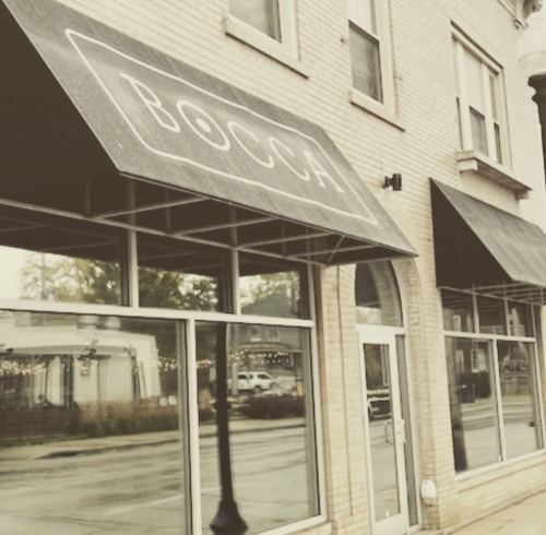 A storefront with the sign "BOCCA" and large awnings, featuring large windows and a sidewalk view.