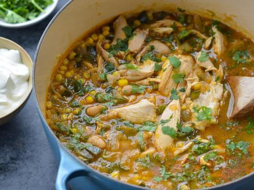 A pot of chicken soup with corn and herbs, garnished with cilantro, alongside bowls of sour cream and greens.