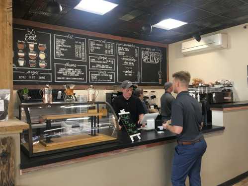 A coffee shop counter with a menu board, a barista serving a customer, and display cases with pastries.