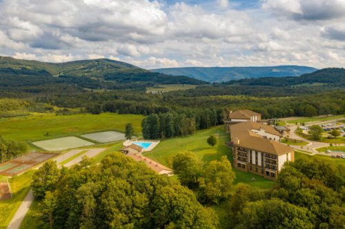 Aerial view of a resort surrounded by green fields and mountains, featuring a pool and tennis courts.