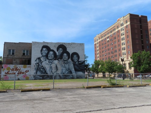 A mural of five people smiling on a building, with a nearby brick building and a fenced area in the foreground.