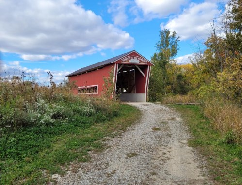 A red covered bridge stands at the end of a gravel path, surrounded by greenery and under a blue sky with clouds.