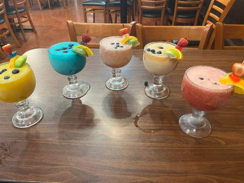 Five colorful frozen cocktails in glasses, garnished with fruit and berries, arranged on a wooden table.