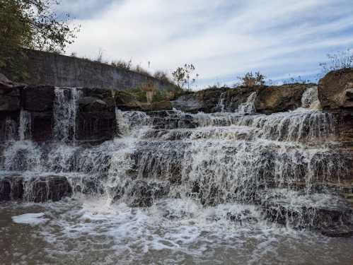 A cascading waterfall flows over rocky ledges, surrounded by greenery and a cloudy sky.