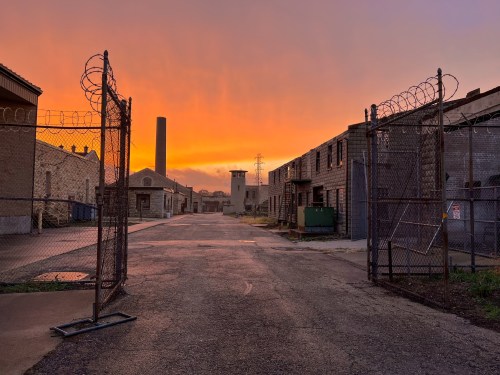 A deserted industrial area at sunset, featuring barbed wire fences and old buildings against a colorful sky.