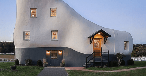 A whimsical, curved house with large windows, a welcoming entrance, and a grassy lawn at dusk.