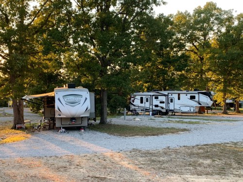Two RVs parked under trees at a campsite, with gravel paths and a warm sunset glow in the background.