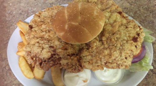 A large fried chicken breast on a bun, served with lettuce, onion, fries, and two dipping sauces on a white plate.