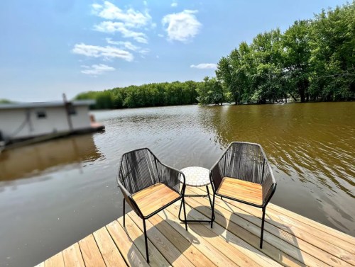 Two chairs and a small table on a wooden deck overlooking a calm river surrounded by trees under a blue sky.