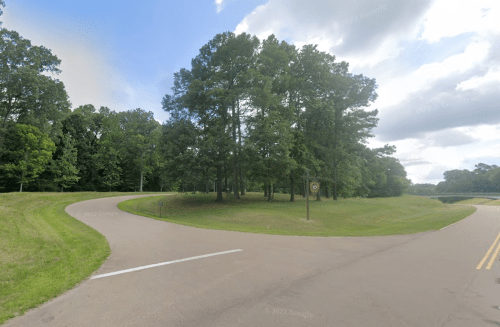 A winding road curves around a grassy area with trees under a partly cloudy sky.