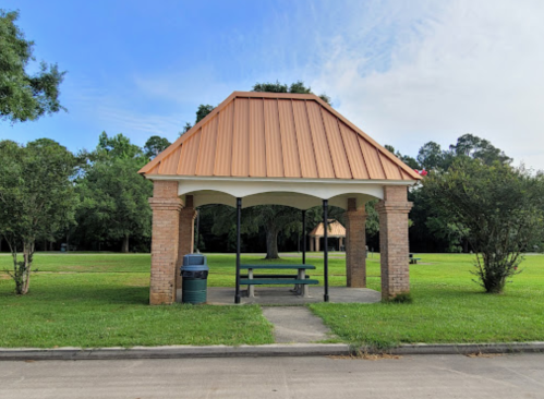 A small gazebo with a copper roof and brick columns in a grassy park area, with a picnic table underneath.