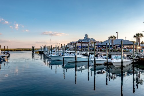 A serene marina at sunset, featuring boats docked along wooden pilings and a lively waterfront restaurant.