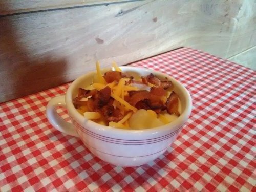 A bowl of macaroni and cheese topped with crispy bacon and shredded cheese, set on a red and white checkered tablecloth.