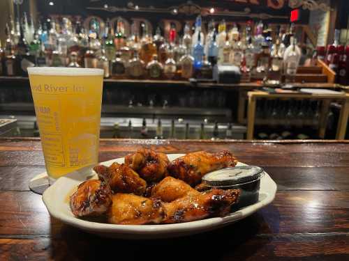 A plate of glazed chicken wings with a side of dipping sauce, next to a glass of beer, set against a bar backdrop.