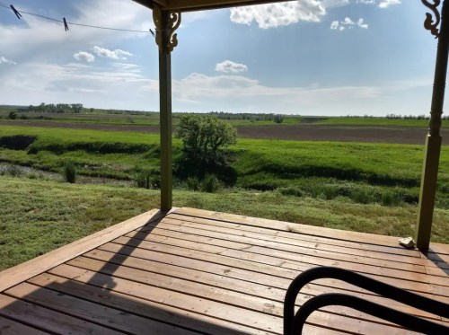A wooden porch overlooks a lush green field under a bright blue sky with scattered clouds.