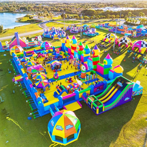 Aerial view of a colorful inflatable bounce park with various slides, castles, and play areas on a sunny day.