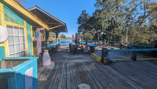 A colorful outdoor dining area with wooden tables, chairs, and a pool in the background under a clear blue sky.