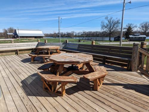A wooden deck with a large octagonal table and benches, surrounded by green fields and a clear blue sky.
