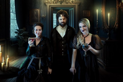 Three elegantly dressed individuals stand in a dimly lit, vintage room, each holding a drink, with candles flickering nearby.