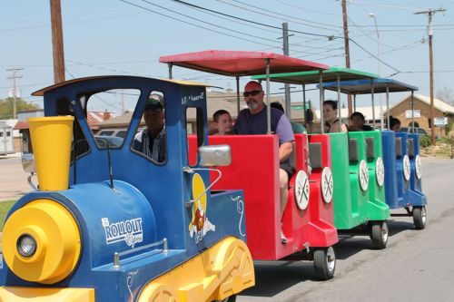 A colorful miniature train with passengers rides along a street, surrounded by power lines and buildings.