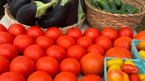 A variety of fresh vegetables, including red tomatoes, eggplants, and green jalapeños, displayed in baskets.
