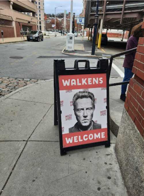 A sidewalk sign featuring a black-and-white portrait of a man with the text "WALKENS WELCOME" in bold red letters.