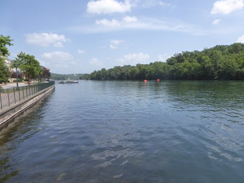 A calm river scene with green trees lining the banks and a clear blue sky, featuring a dock and red buoys in the water.
