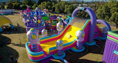 A colorful inflatable water slide at a fun park, surrounded by various other inflatable attractions and trees.