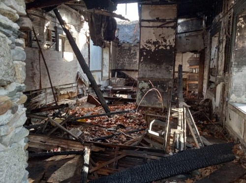 Interior of a burned building, with charred debris, fallen leaves, and damaged walls, creating a desolate atmosphere.