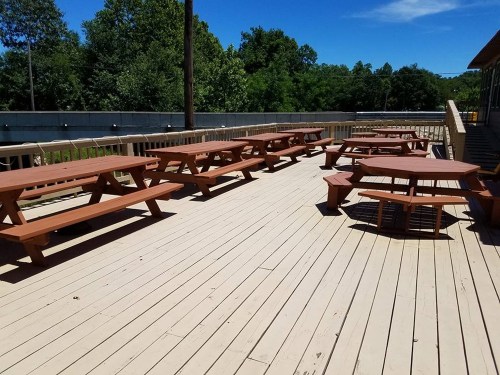 A spacious outdoor deck with several wooden picnic tables under a clear blue sky and green trees in the background.