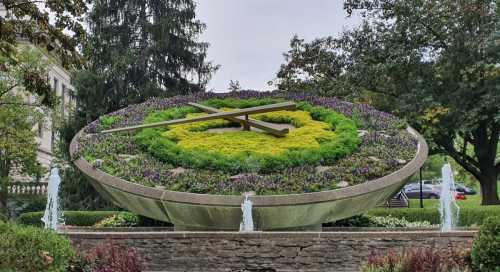 A large floral clock surrounded by greenery and fountains, set in a park-like area with trees in the background.