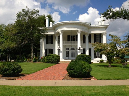 A grand white mansion with columns, surrounded by green lawns and trees, under a partly cloudy sky.
