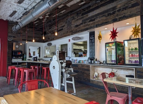 A cozy café interior with wooden beams, red chairs, and a counter displaying drinks and snacks. Brightly lit and inviting.