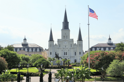 A historic building with spires, surrounded by greenery and a flag, set against a clear blue sky.