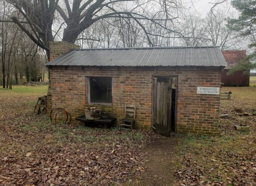 A small, rustic brick building with a metal roof, surrounded by trees and fallen leaves.