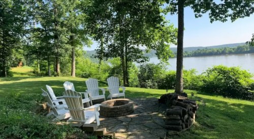 A serene lakeside scene featuring white Adirondack chairs around a stone fire pit, surrounded by lush greenery.