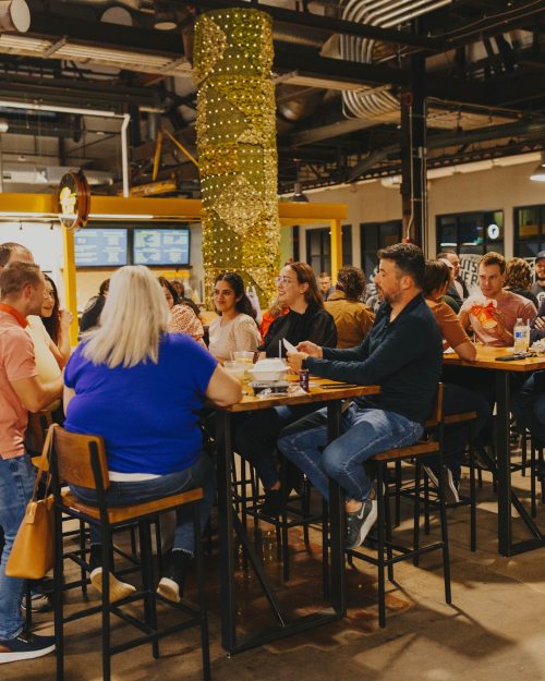 A lively restaurant scene with people seated at high tables, enjoying food and drinks in a vibrant atmosphere.