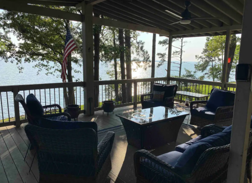 A cozy porch with wicker furniture and a view of a lake, surrounded by trees and an American flag.