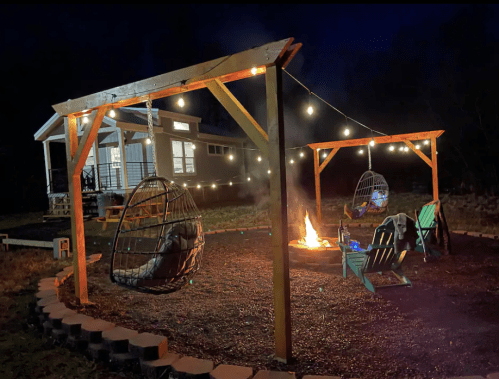 Cozy outdoor space at night with hanging chairs, a fire pit, and string lights illuminating a home in the background.