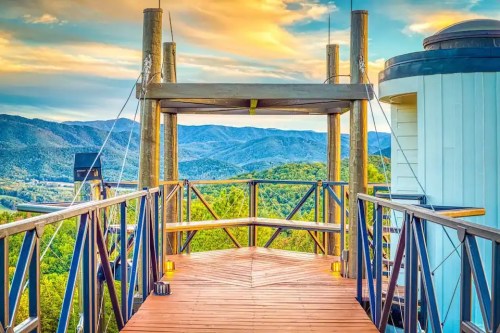 A wooden deck with a railing overlooks lush green mountains under a colorful sky at sunset.