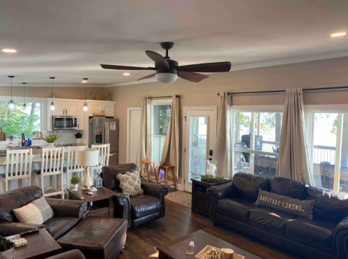 Cozy living room with leather sofas, a ceiling fan, and large windows overlooking a lake. Modern kitchen in the background.