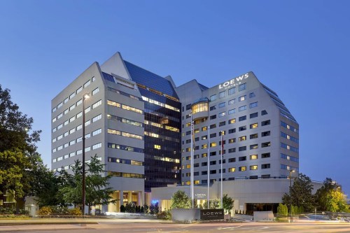 Modern hotel building at dusk, featuring large windows and a prominent sign for Loews. Trees and street visible in foreground.