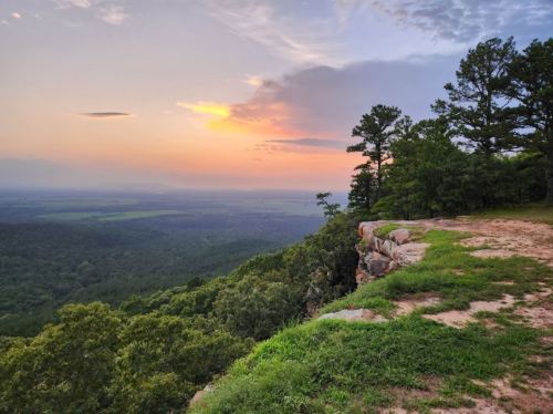 A scenic view of a sunset over a lush green valley, with rocky cliffs and trees in the foreground.