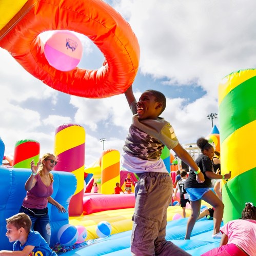 A joyful child jumps to catch a beach ball in a colorful inflatable play area, surrounded by other kids and adults.