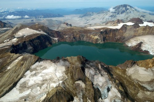 Aerial view of a volcanic crater lake surrounded by rugged terrain and snow-capped mountains under a clear sky.