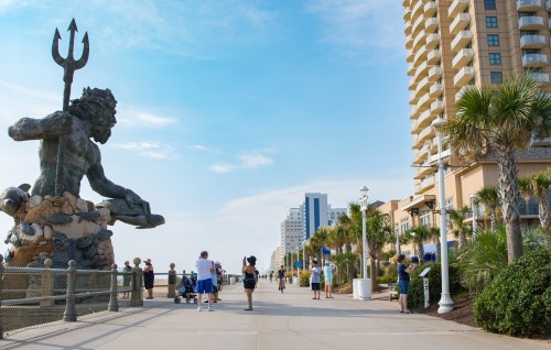 A coastal boardwalk with a large statue of a sea god, surrounded by palm trees and people enjoying the sunny day.